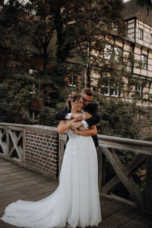 Bride and groom embracing on wooden bridge surrounded by lush greenery.