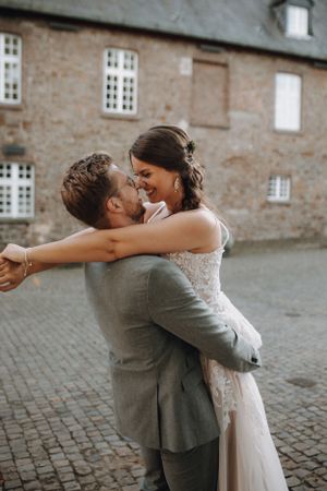 Groom lifting and embracing laughing bride in a lace dress outside a stone building.