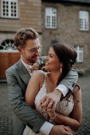 Groom in grey suit embracing bride in lace dress while smiling at each other.