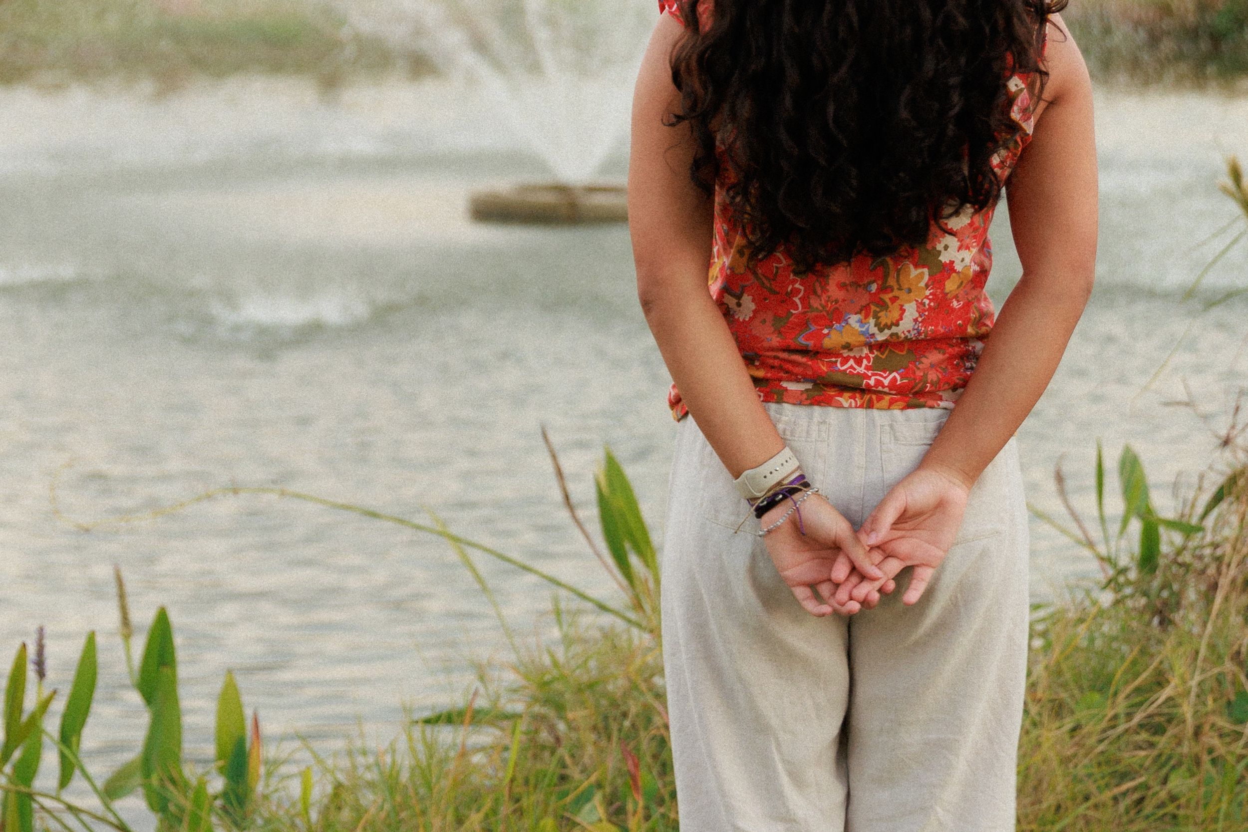 Person in floral top and white pants standing by water.