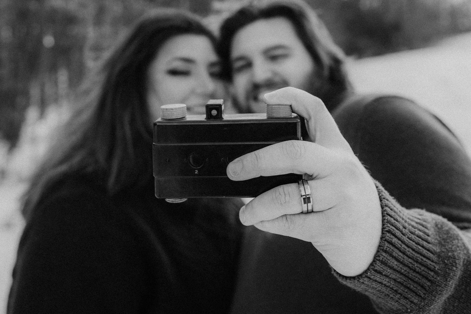 Close-up black and white shot of hands holding vintage camera with couple blurred in background.