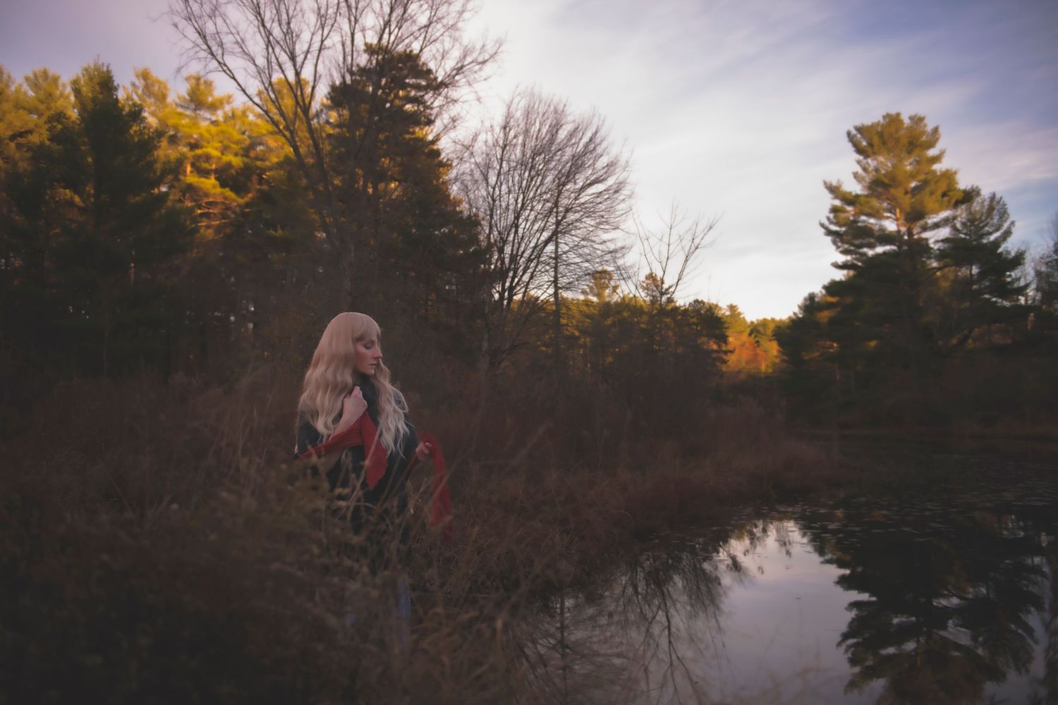 Figure in dark clothing stands near a reflective pond at dusk with autumn trees in background.