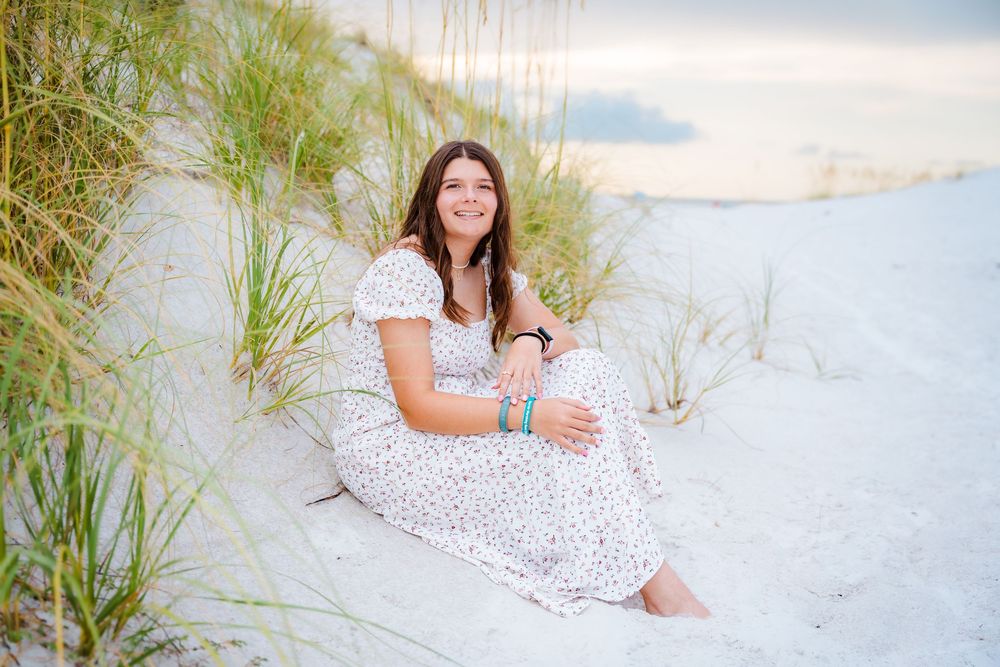 Young woman in white floral dress sitting on white sand dunes with sea grass, smiling at beach sunset.