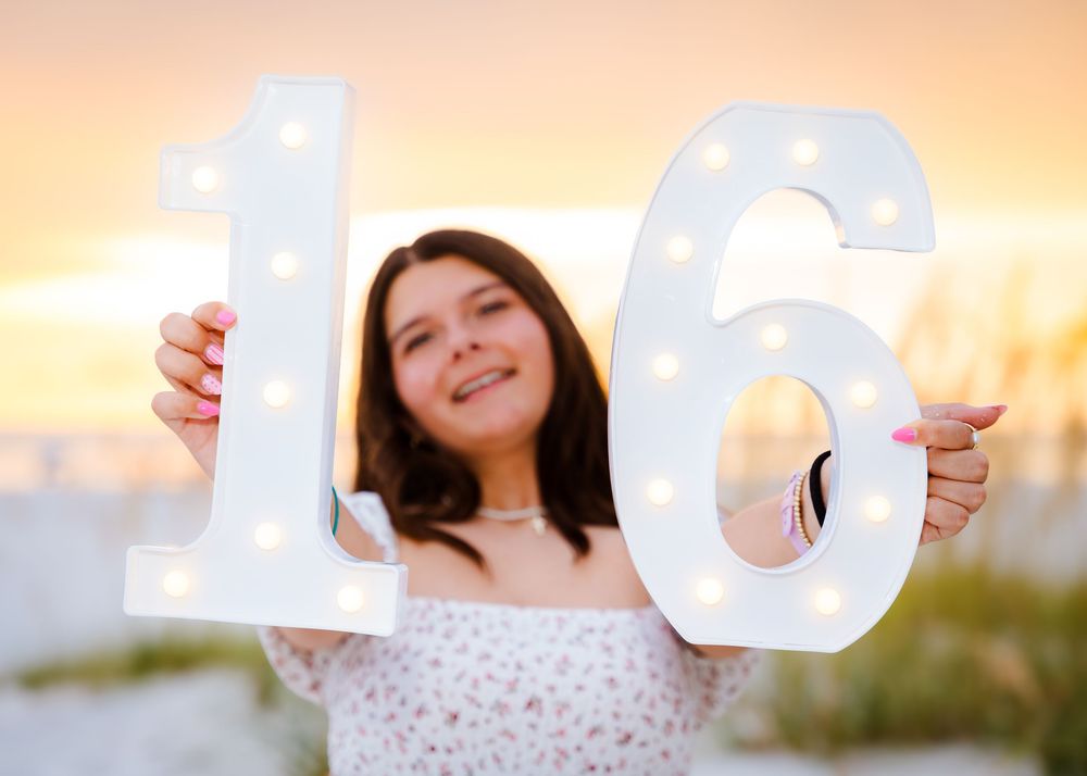 Teen girl smiling and holding illuminated white marquee number 16 signs at golden hour beach sunset.