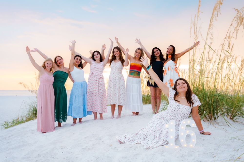 Group of girls celebrating a 16th birthday on a white sand beach at sunset, wearing colorful dresses.