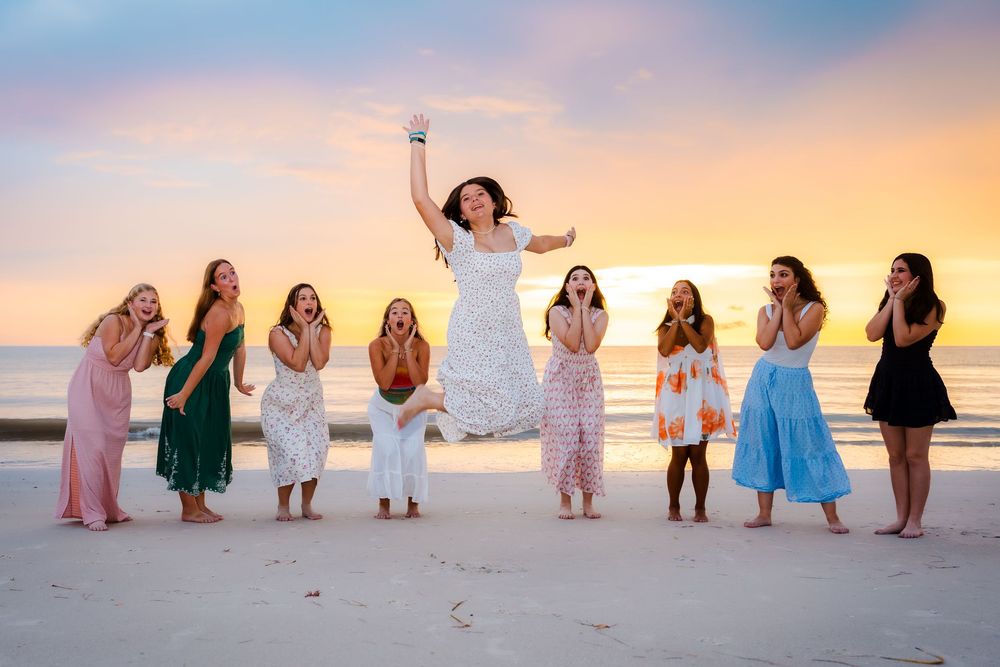 Bride jumps joyfully on beach at sunset surrounded by bridesmaids in colorful dresses during bouquet toss.