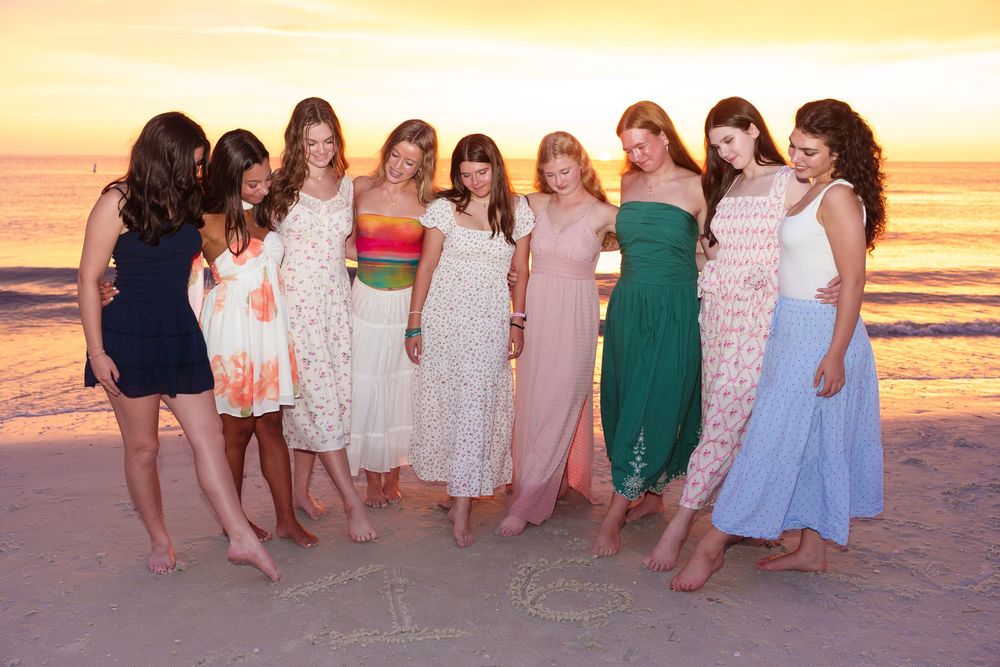 Group of nine women in colorful dresses posing together on a beach at sunset.