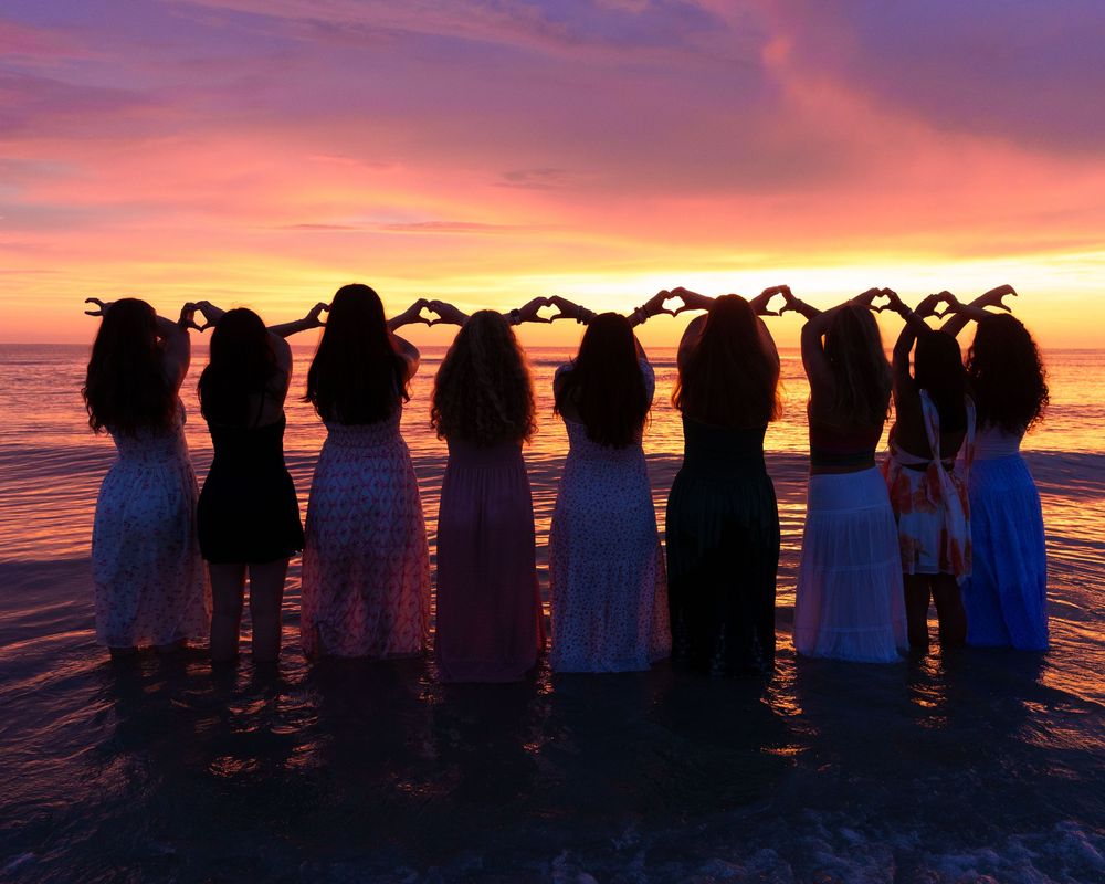 Eight girls standing in ocean water at sunset, forming heart shapes with their hands against a vibrant pink and orange sky.