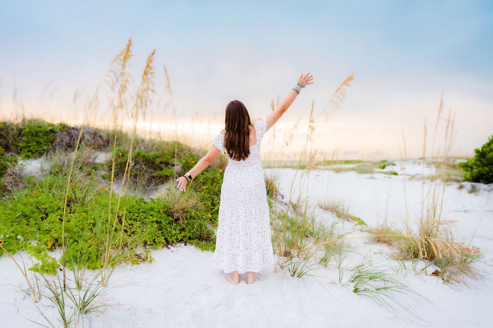 Woman in white dress standing on white sand dunes with arms raised, surrounded by sea oats at sunset.