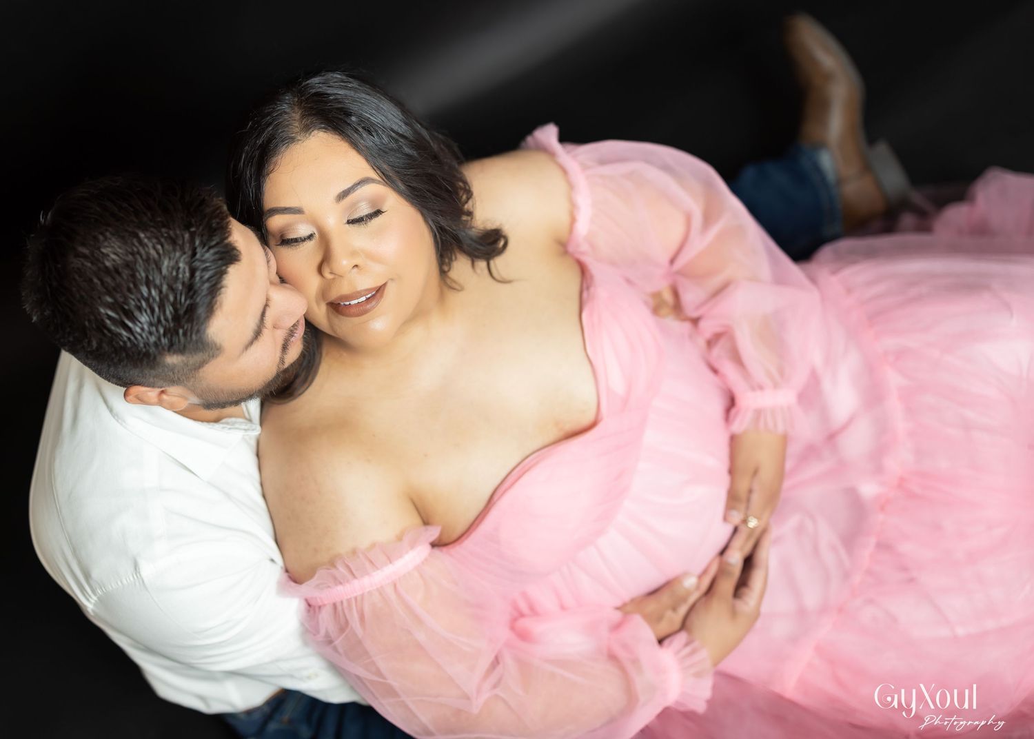 A romantic maternity photo featuring a couple embracing in pink flowing fabric against a dark background.