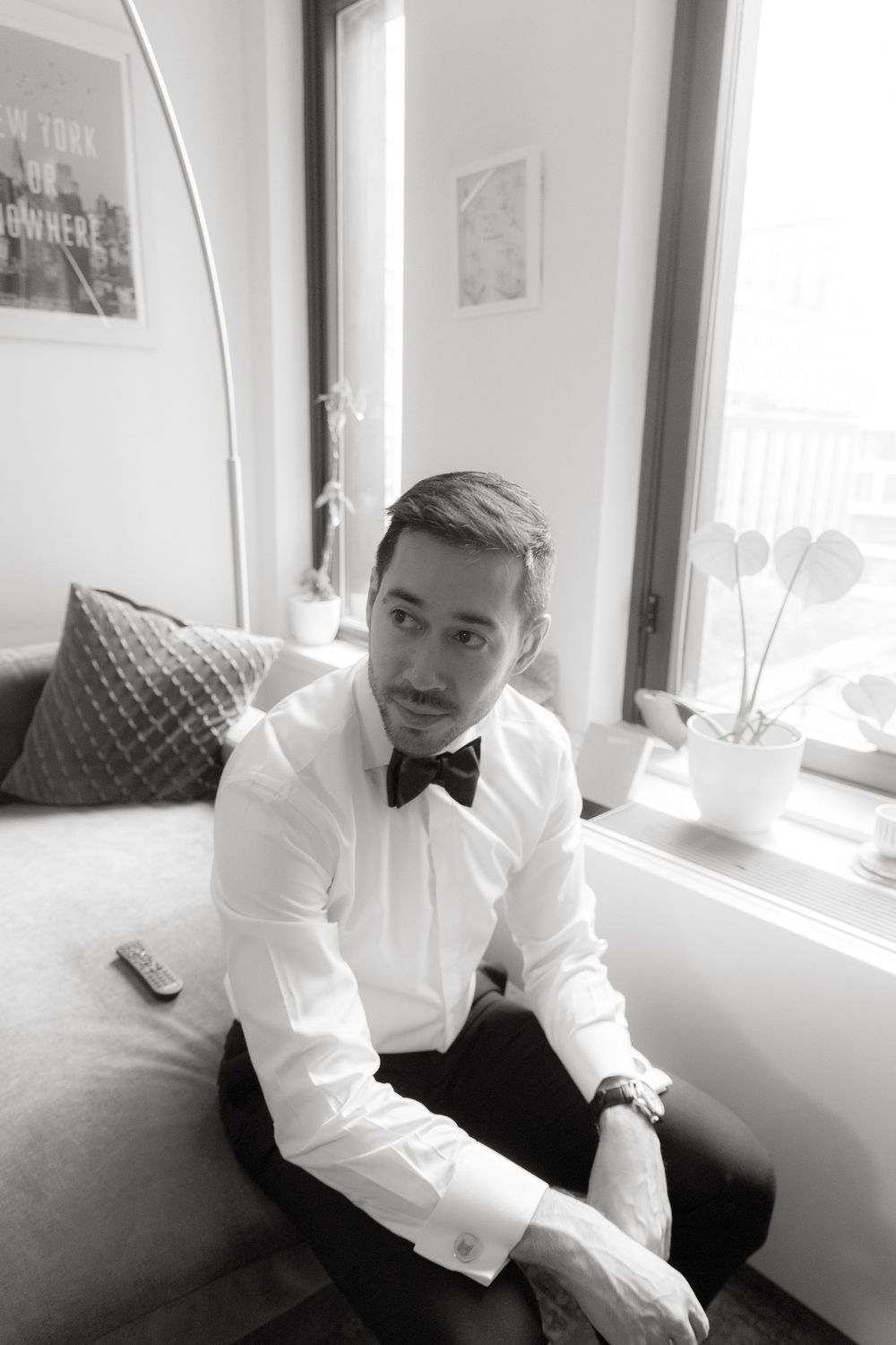 Man in formal white shirt and bow tie gazing pensively by window in black and white portrait series.