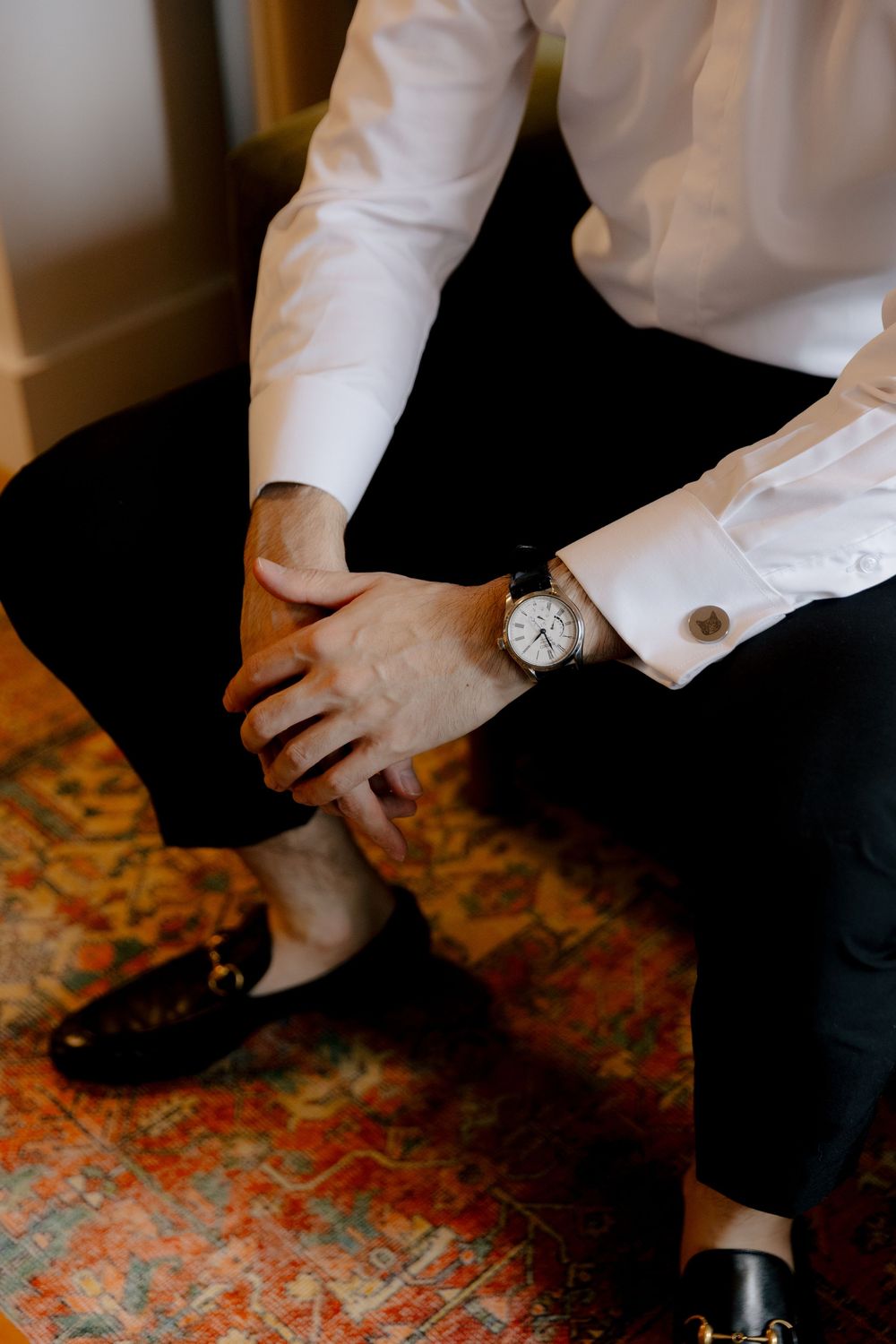 Close-up of groom's formal attire showing watch and cufflinks while seated on patterned carpet.