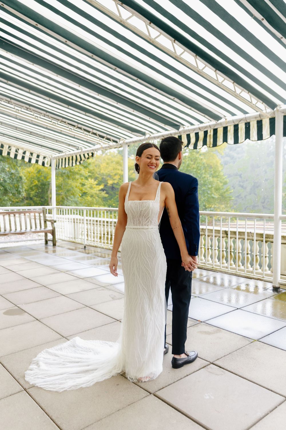 Wedding couple stands on a covered terrace with striped awning and garden views.