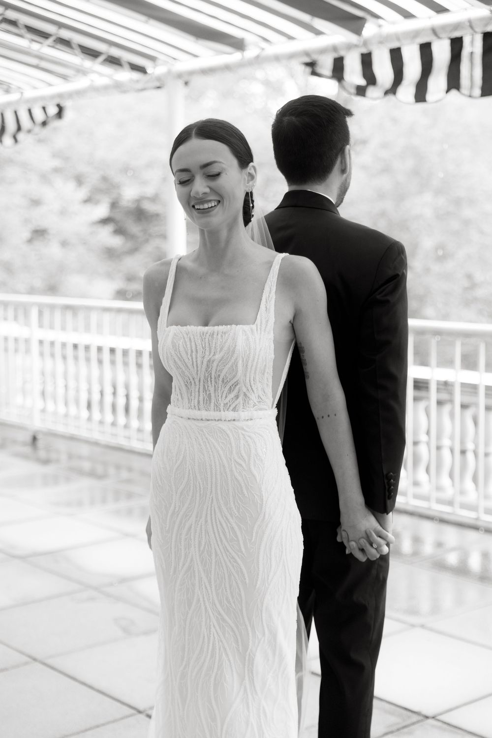 Black and white wedding photo showing a bride in a fitted sleeveless gown holding hands with groom under a striped canopy.