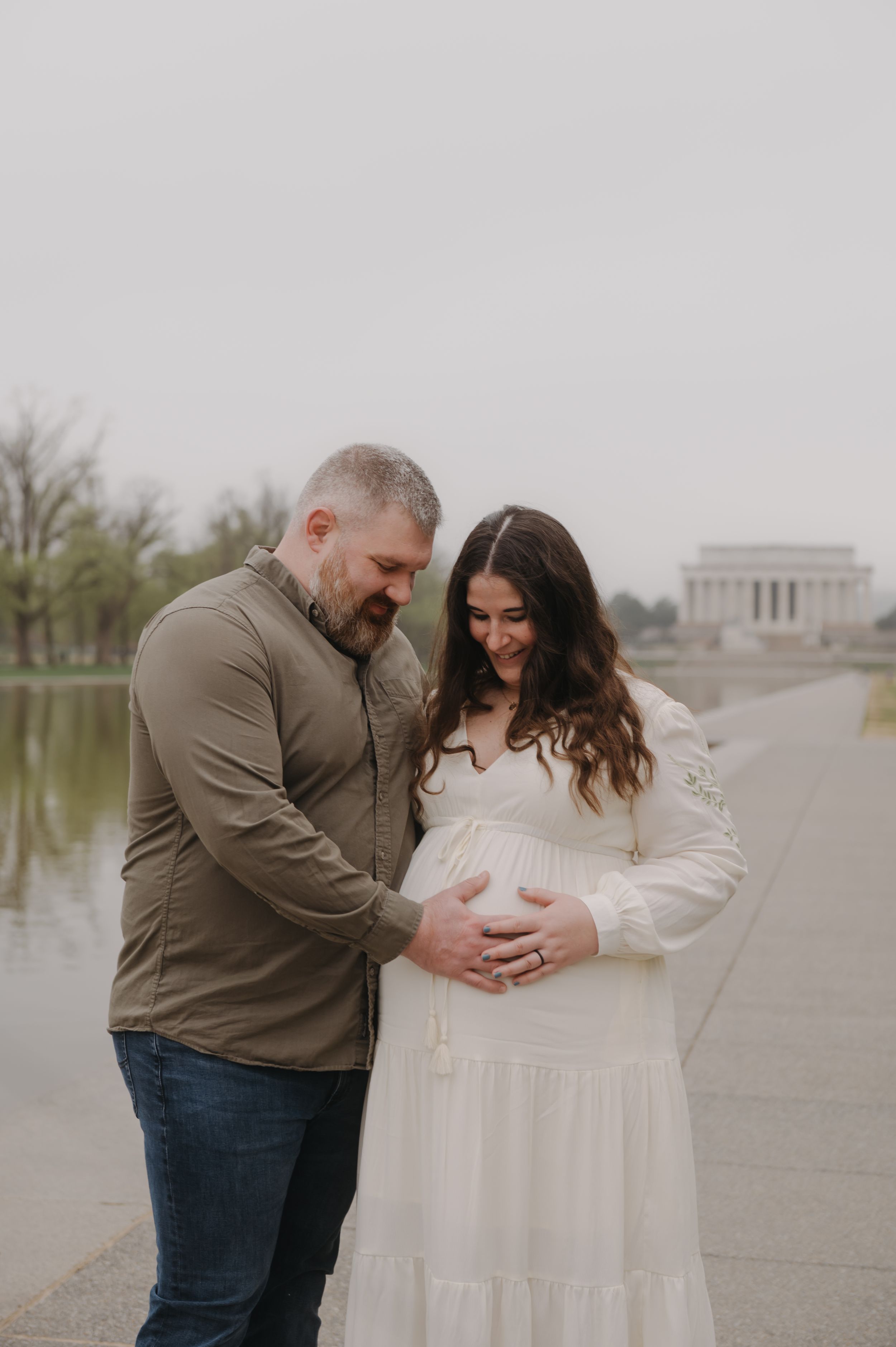 Expectant couple share tender moment near Lincoln Memorial reflecting pool on overcast day in Washington DC.