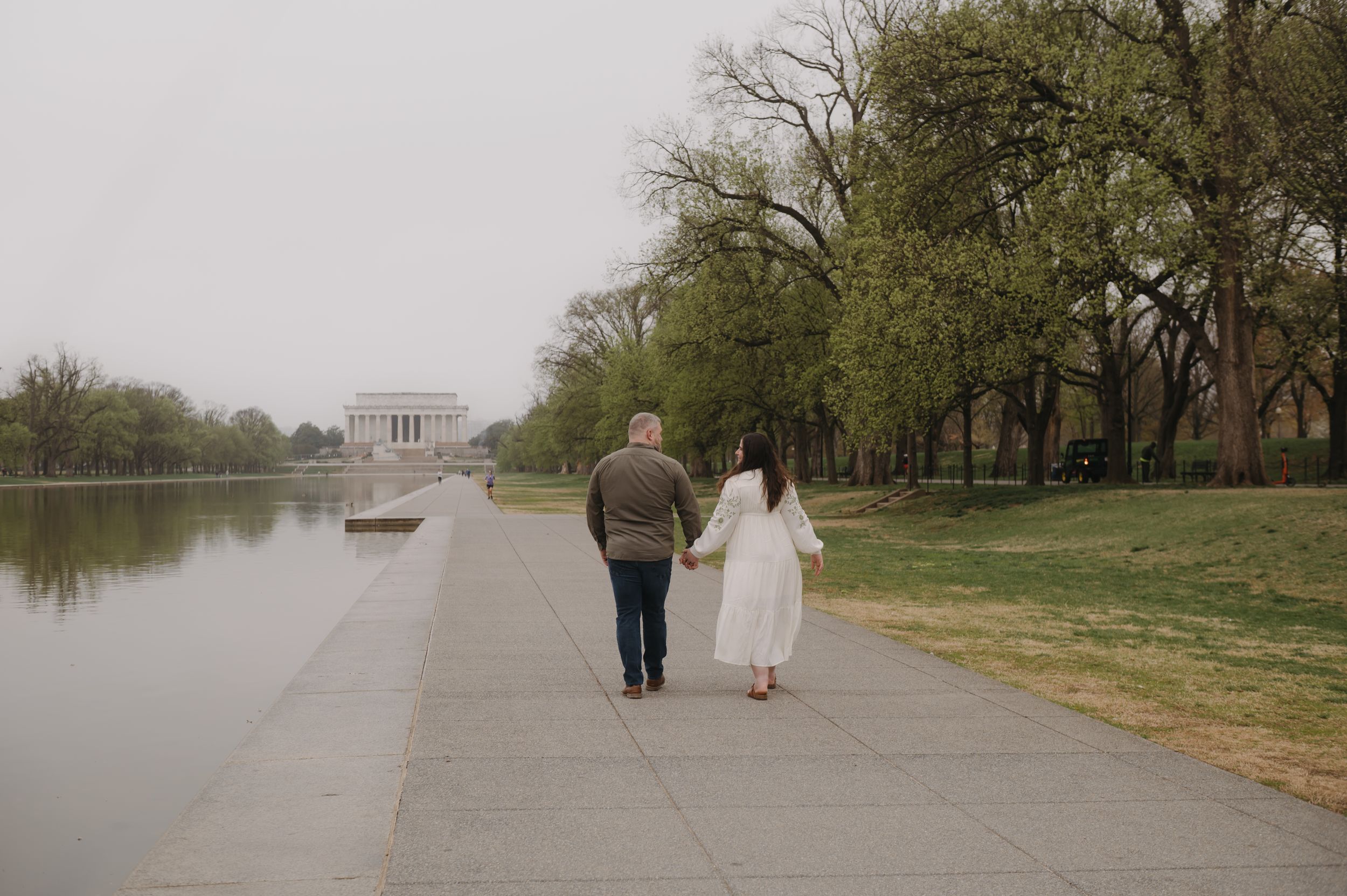 Two people walking on path along reflecting pool towards Lincoln Memorial with trees lining the walkway.