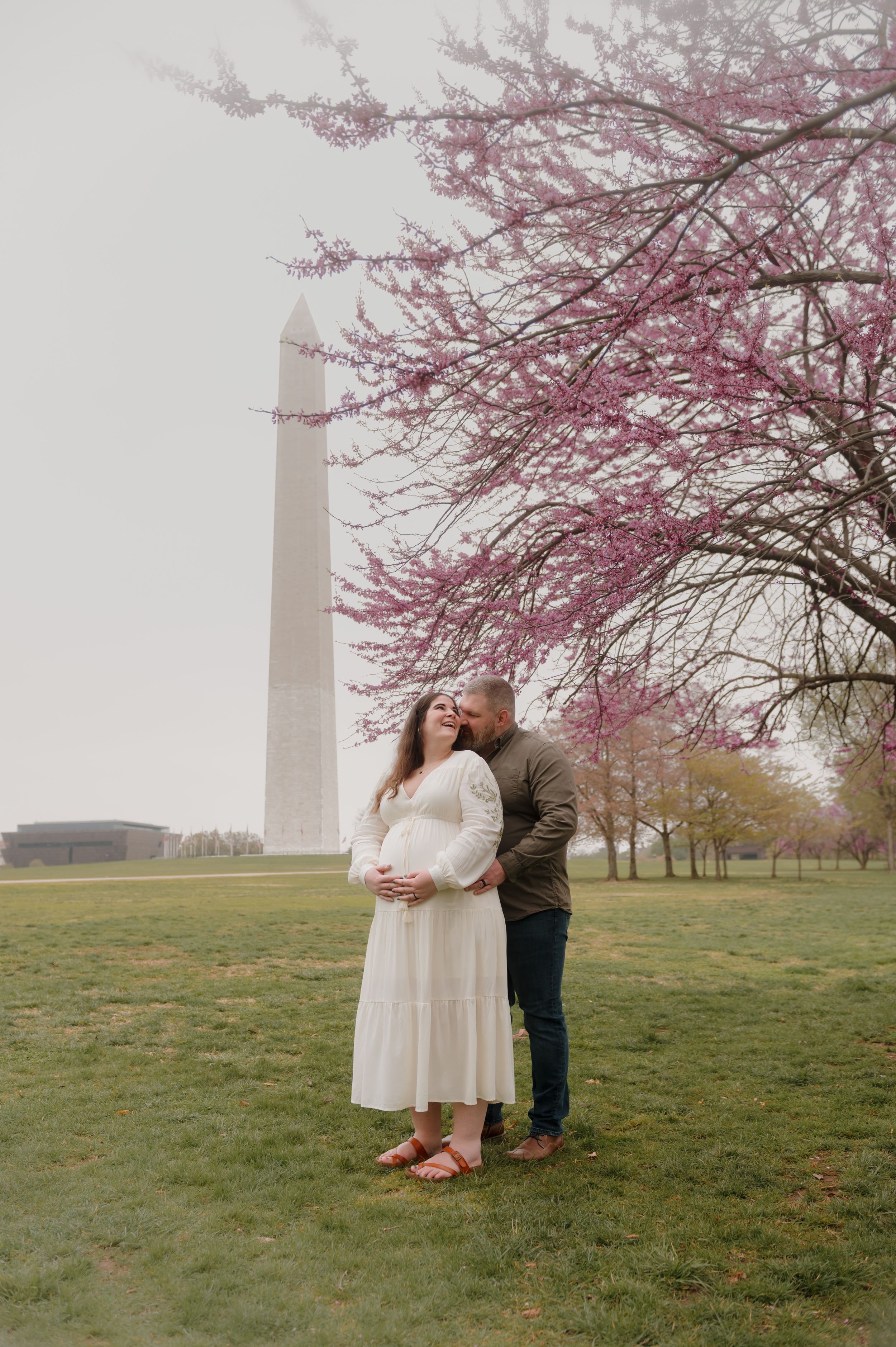 A couple stands beneath blooming cherry blossoms near the Washington Monument on a misty spring day.