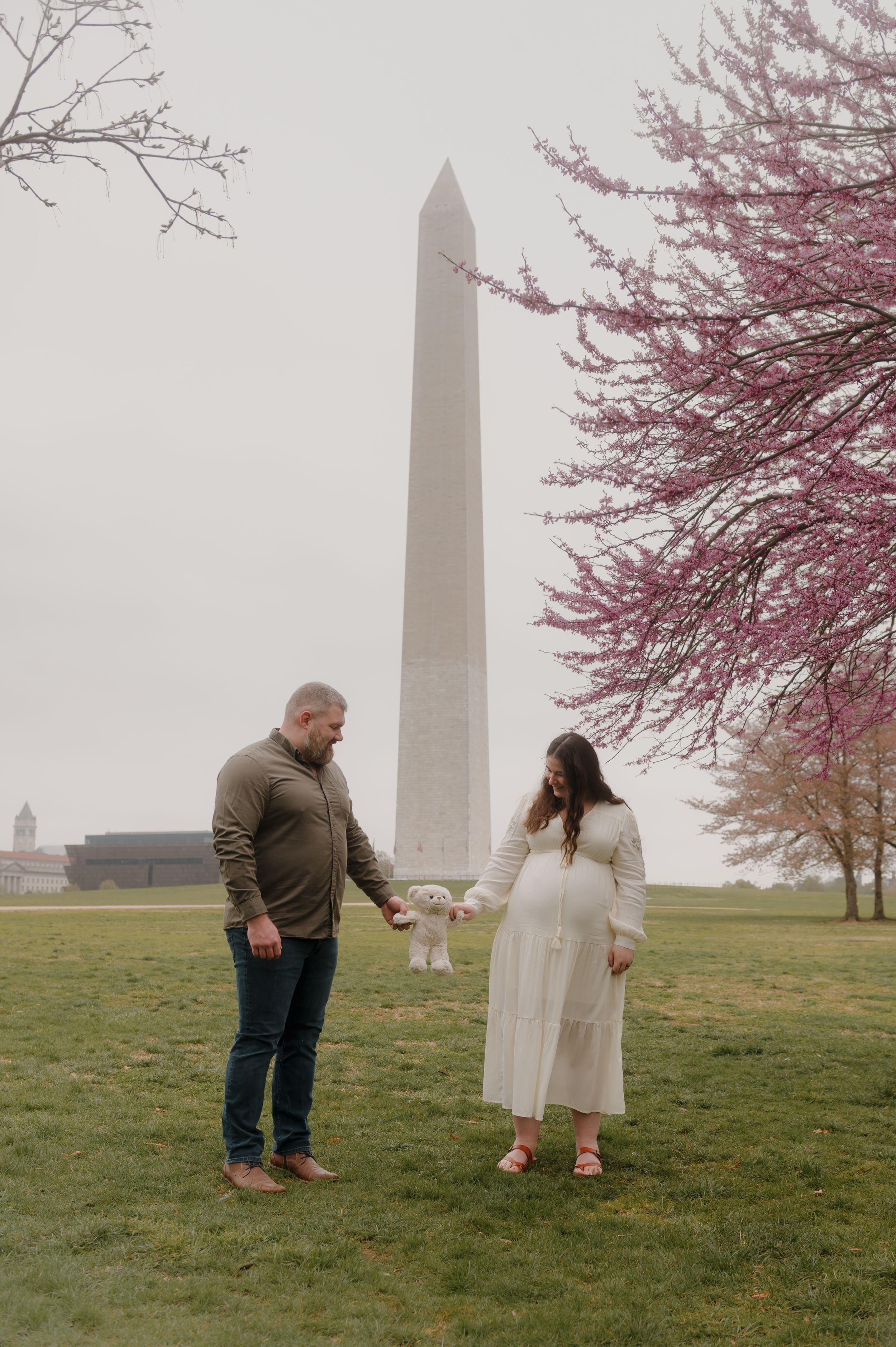 Playful couple poses by Washington Monument while surrounded by pink cherry blossoms.