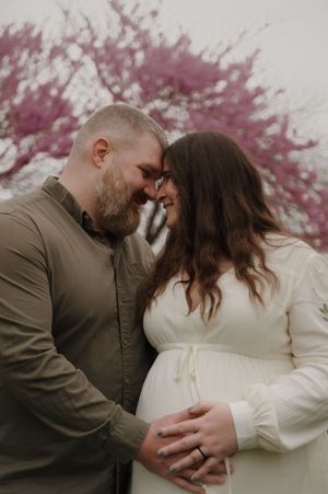 A couple shares an intimate moment under blooming cherry blossom trees during a maternity photoshoot.