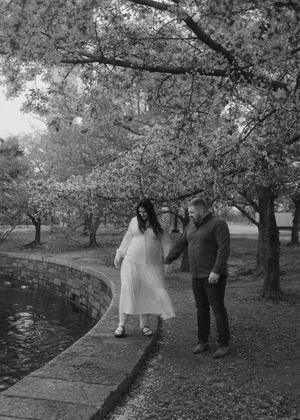 Couple walks along curved path under cherry blossom trees near reflecting pool in monochrome photography.