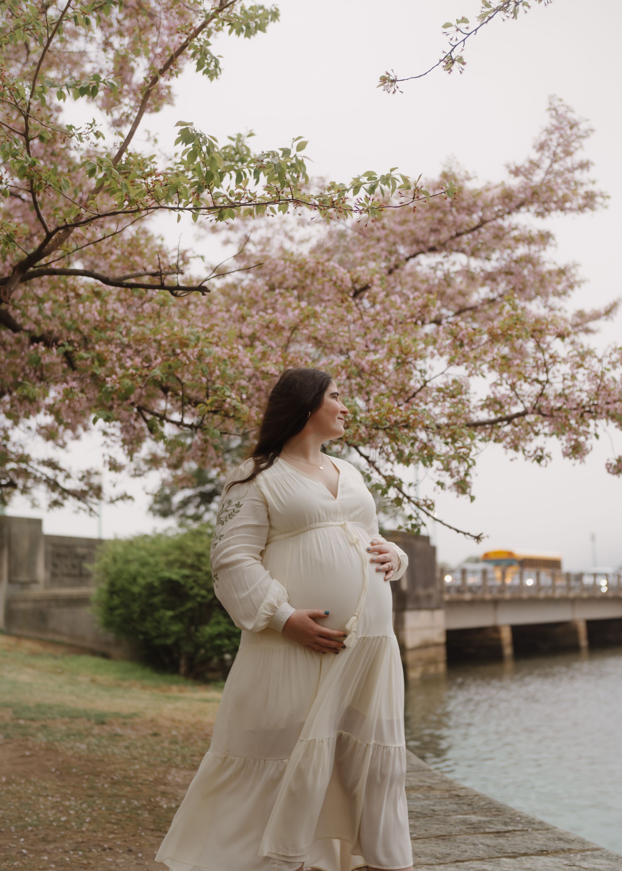 Expecting mother in a white dress poses by cherry blossoms near a waterfront.