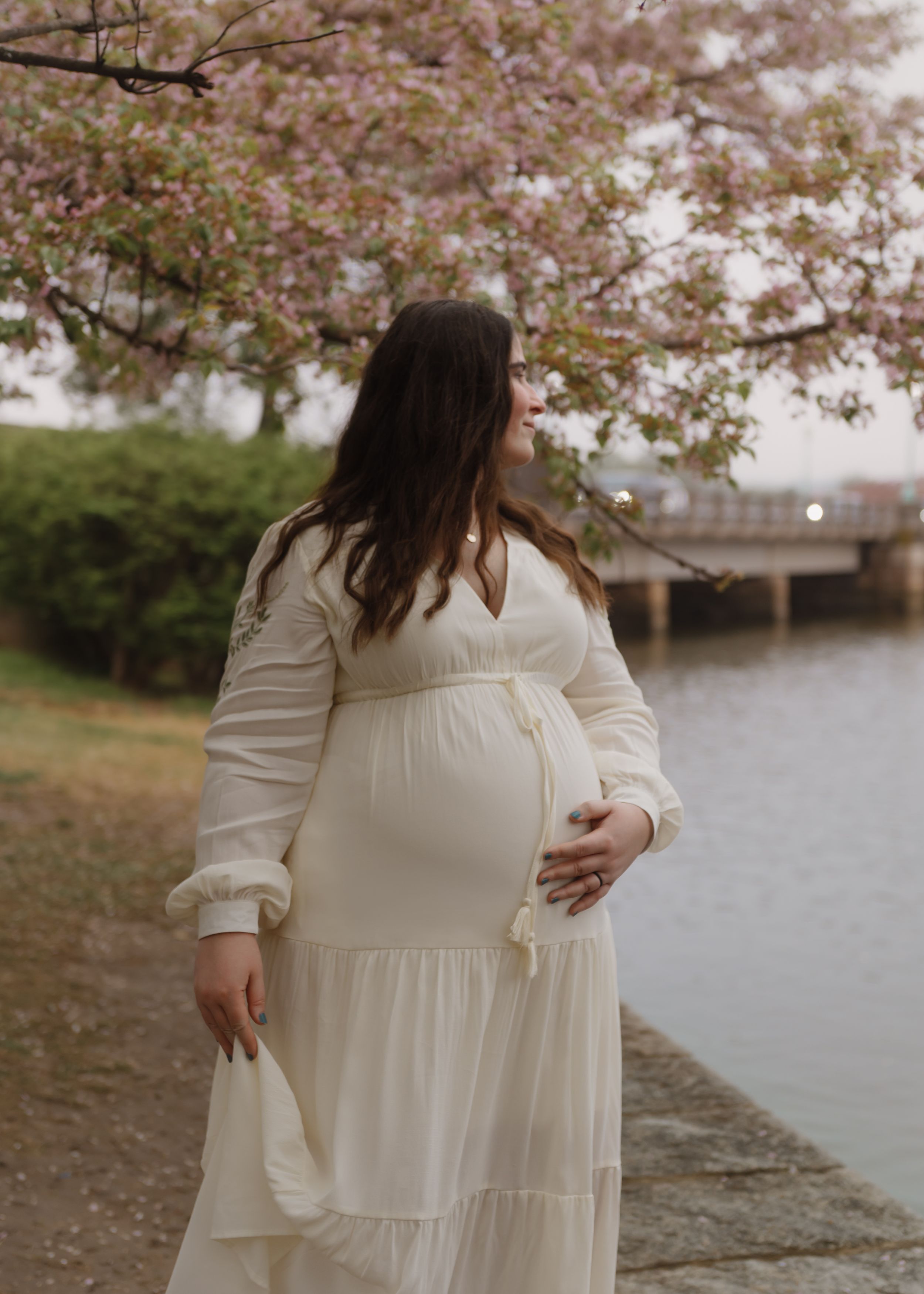 A pregnant woman in a flowing white dress stands near blooming cherry blossoms beside a waterfront.