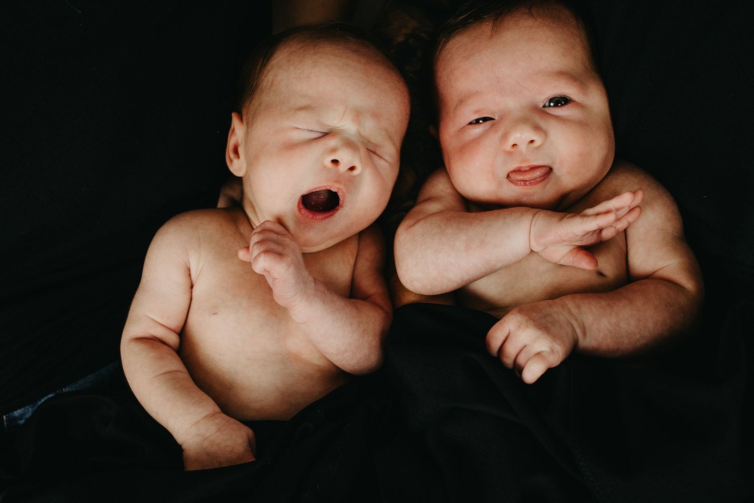 Newborn twins cozied up together laying on black backdrop. One yawns while the other blows raspberries.
