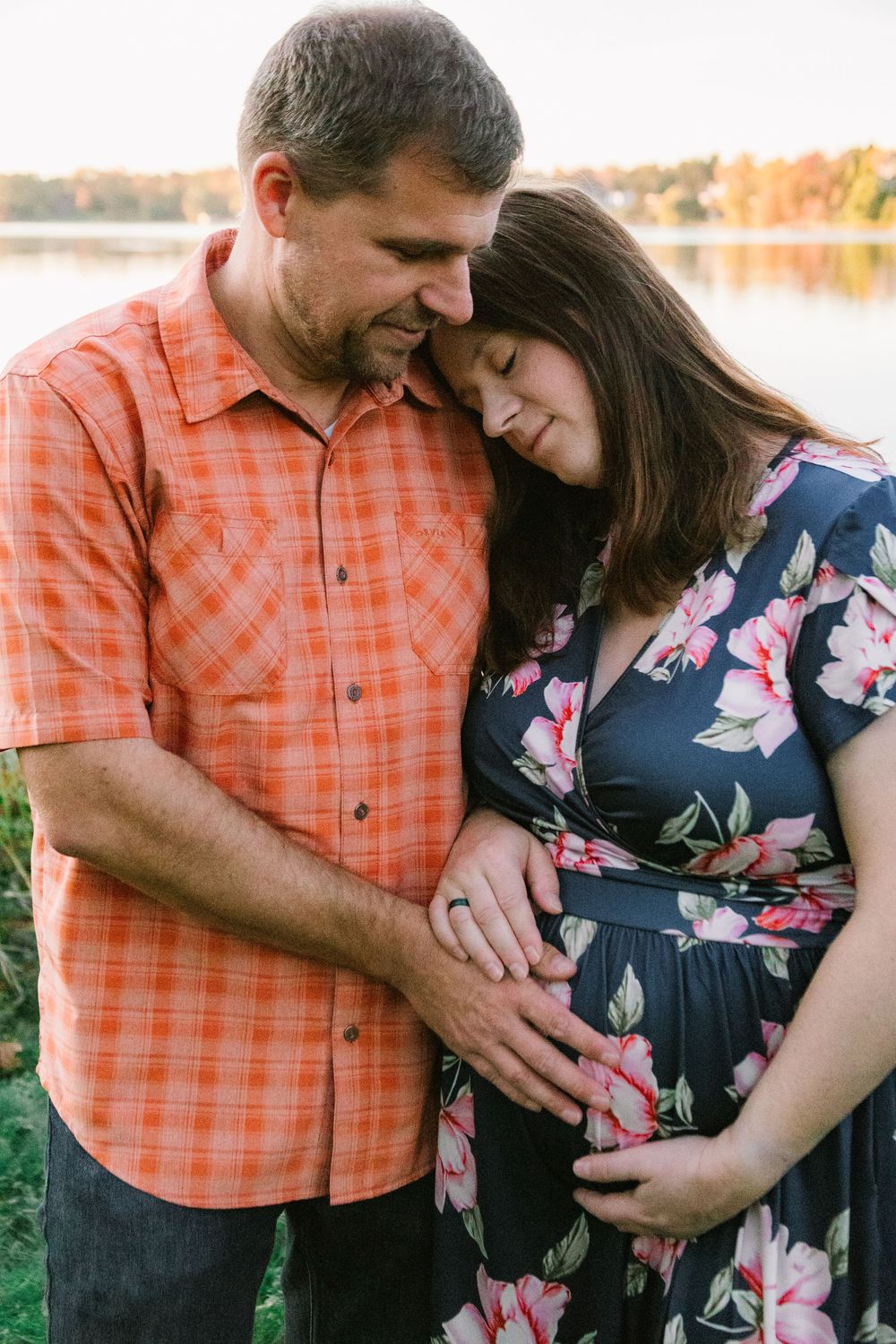 A couple in orange plaid shirt and floral dress share intimate maternity photos by a lake at sunset.