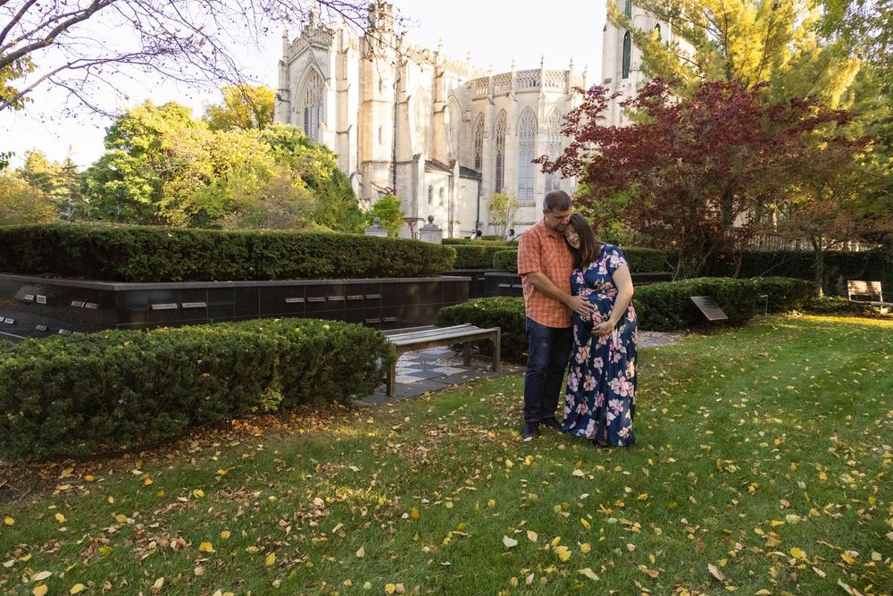 A couple stands together in a garden with autumn foliage and a grand cathedral-style building in the background.