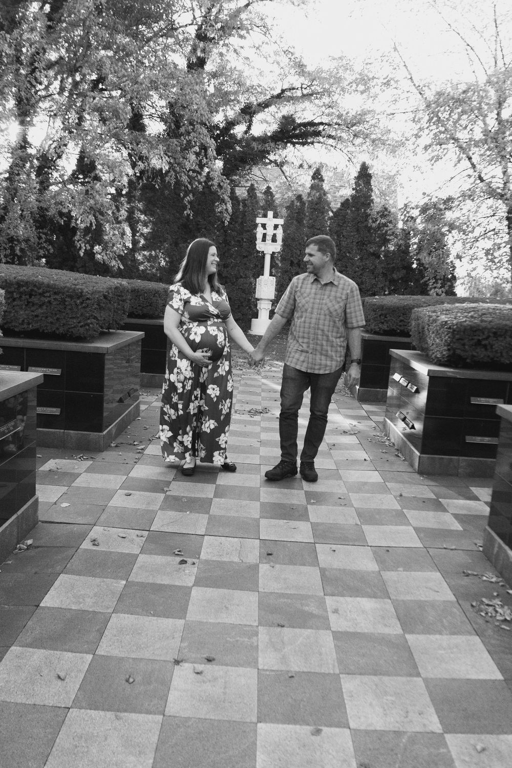 A couple stands on a checkered tile patio as the woman holds up a decorative sign in a floral dress.