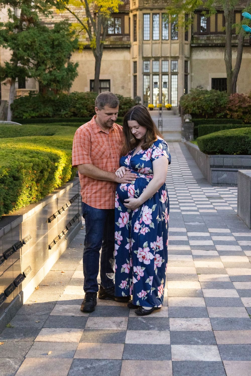 Expectant couple poses on checkered stone pathway surrounded by autumn foliage.