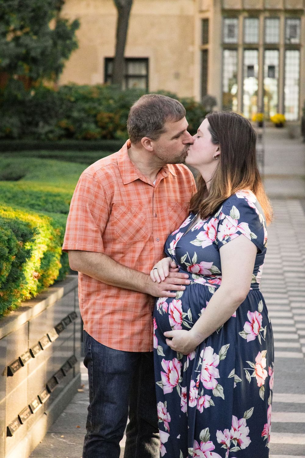 A couple shares romantic kisses while standing on a stone walkway surrounded by landscaping and greenery.