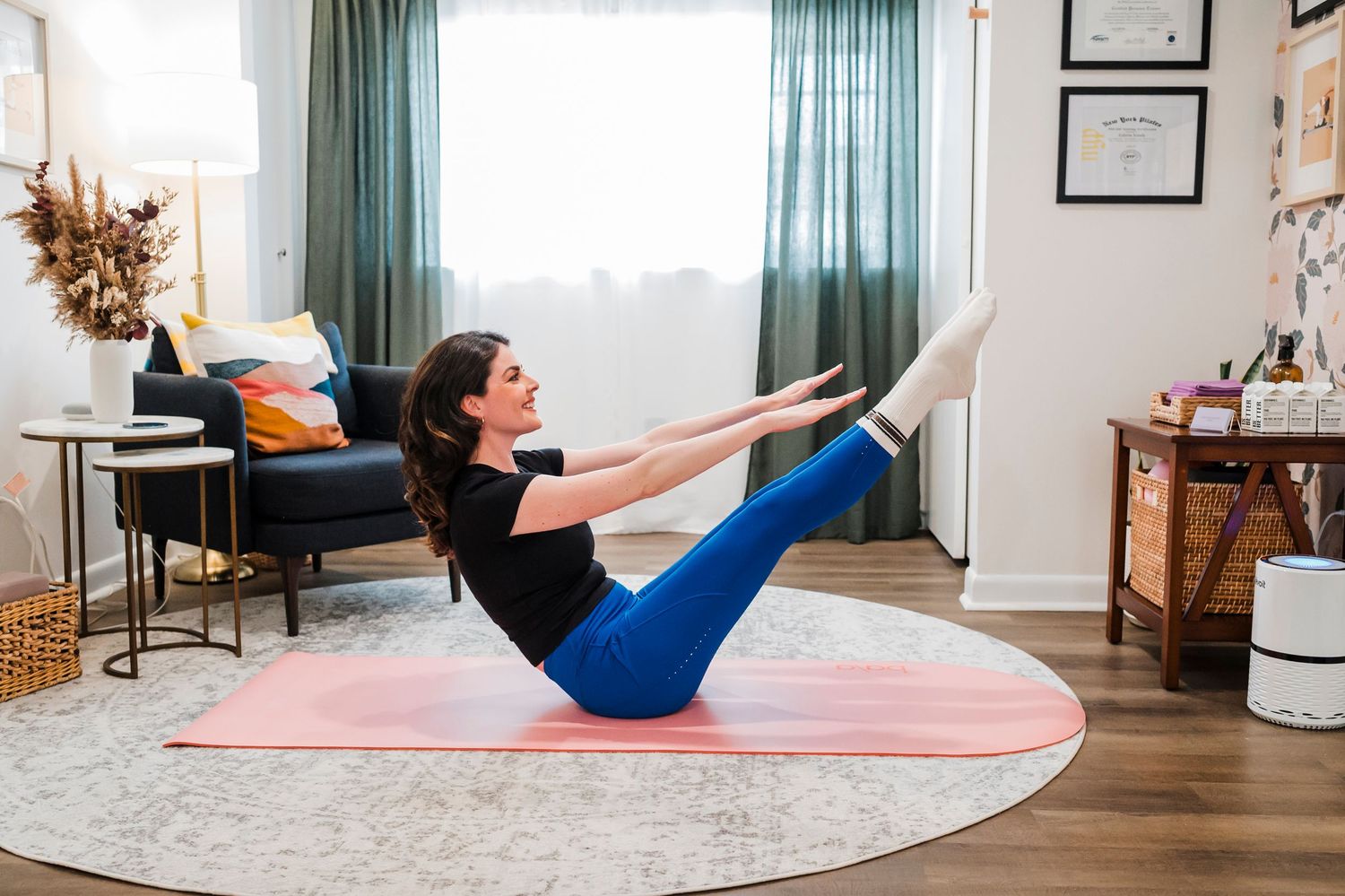 Someone performs a V-sit exercise position on a pink yoga mat in a cozy living room with teal curtains and hardwood floors.