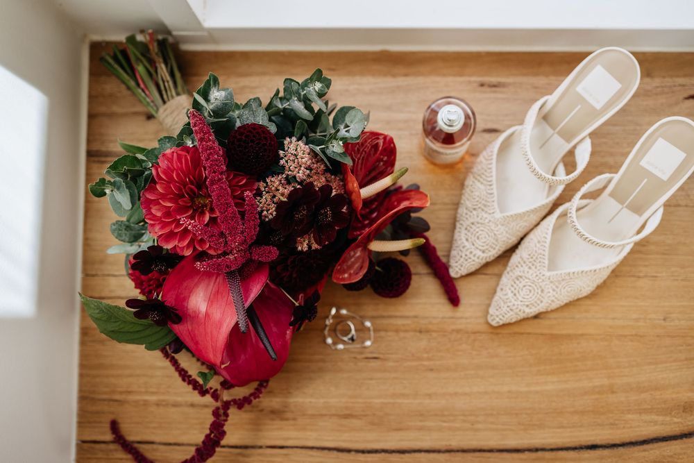 Wedding details with burgundy floral bouquet, white pointed-toe heels and jewelry on wooden surface.
