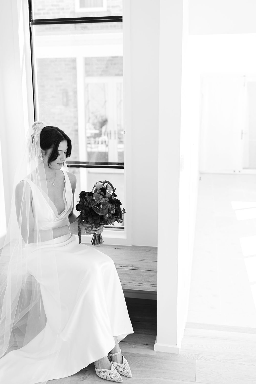 A bride in an elegant white gown sits by a window holding a dark bouquet of flowers in a bright minimal space.