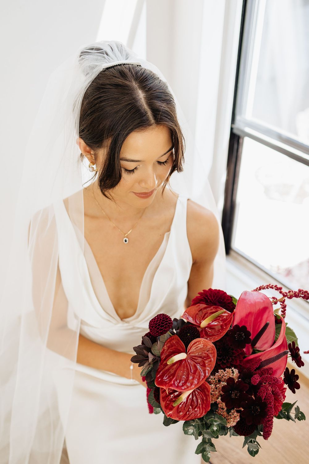 A bride in white holds a stunning red bouquet with anthuriums and roses near a window with soft natural light.
