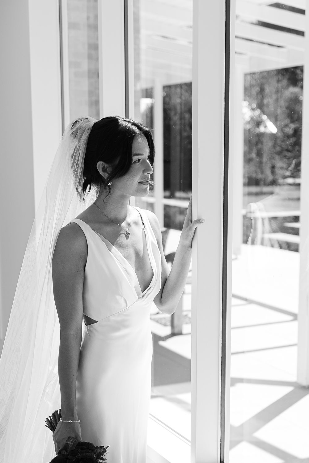 Black and white portrait of a bride in an elegant sleeveless wedding gown and veil standing by a window in natural light.