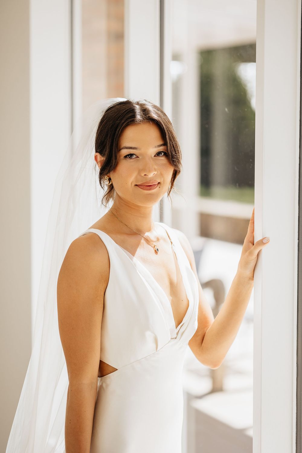 A bride in a white wedding dress poses against a bright window-lit hallway in an elegant setting.