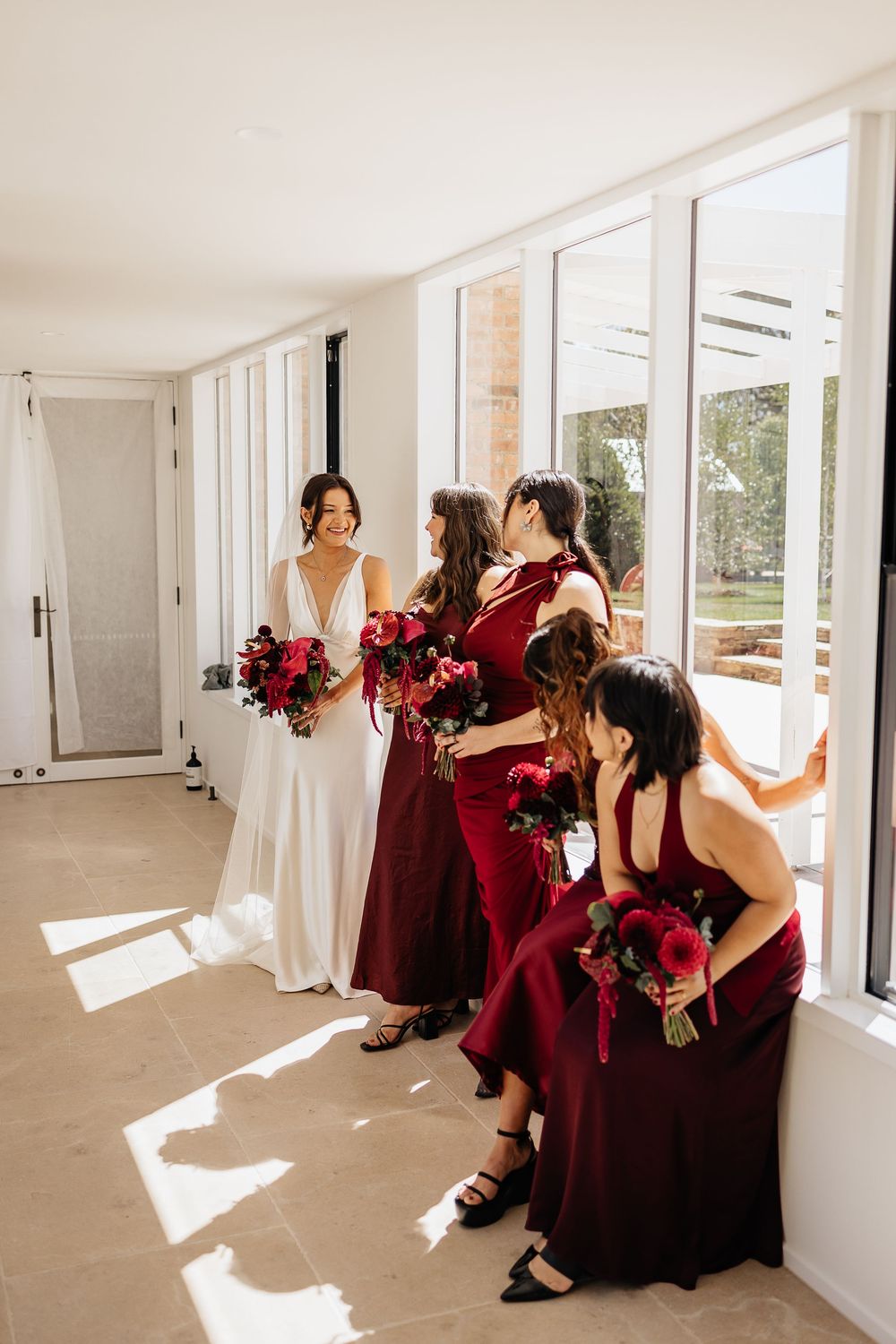 Bridesmaids in burgundy dresses gather by sunlit windows during a wedding celebration.