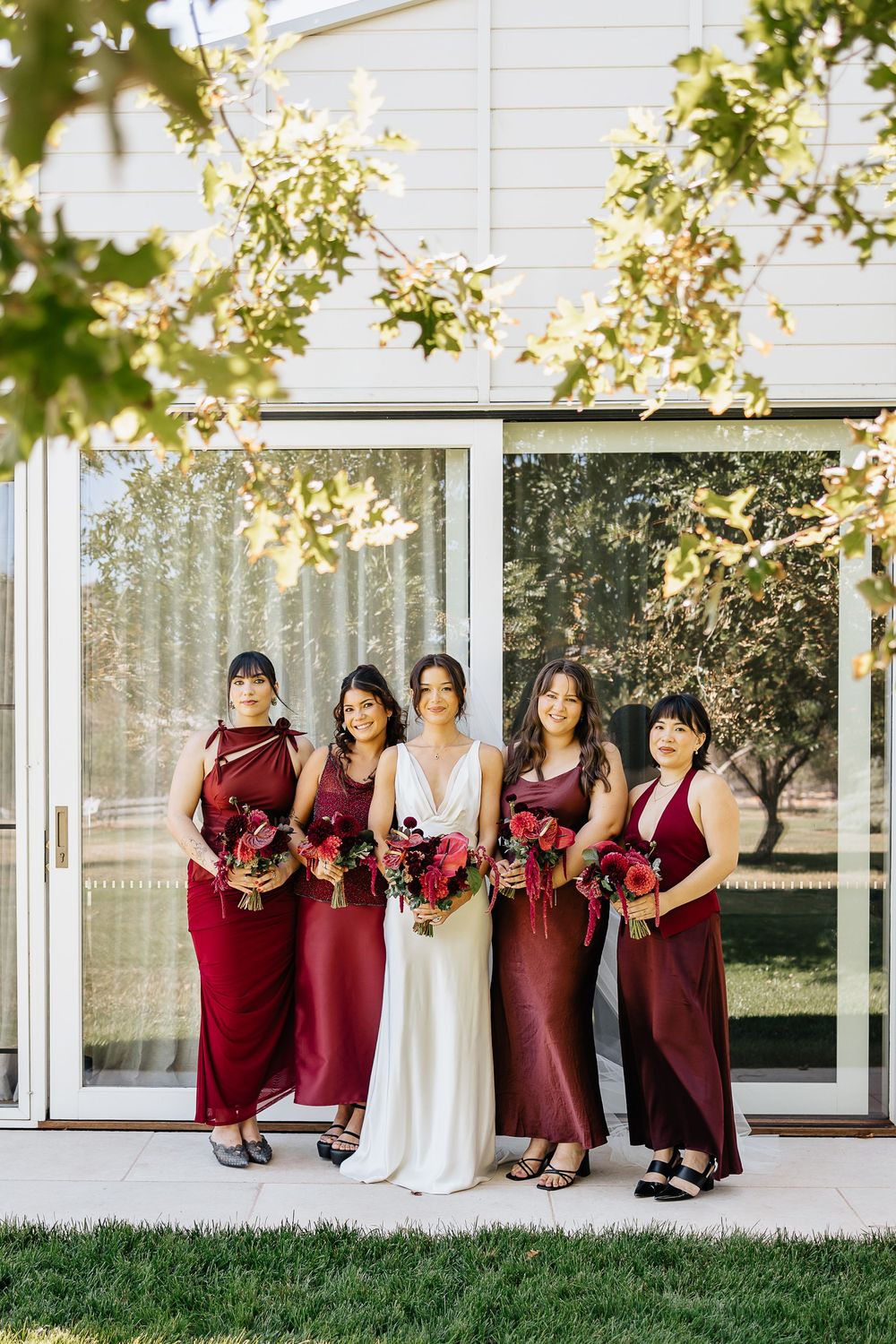Wedding party in burgundy bridesmaid dresses pose together against modern white building exterior.