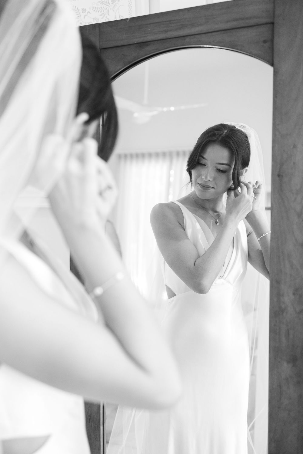 A classic black and white photograph shows a bride looking in a mirror while putting on earrings on her wedding day.