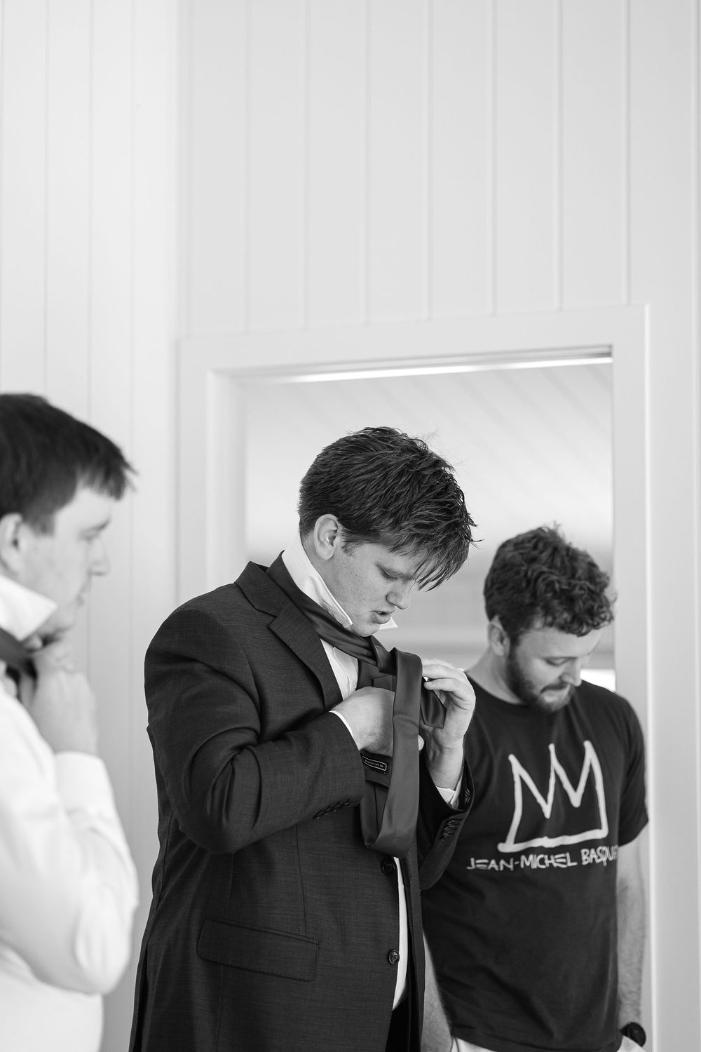 A black and white photograph shows people getting ready, with one adjusting a formal bowtie while wearing a suit.