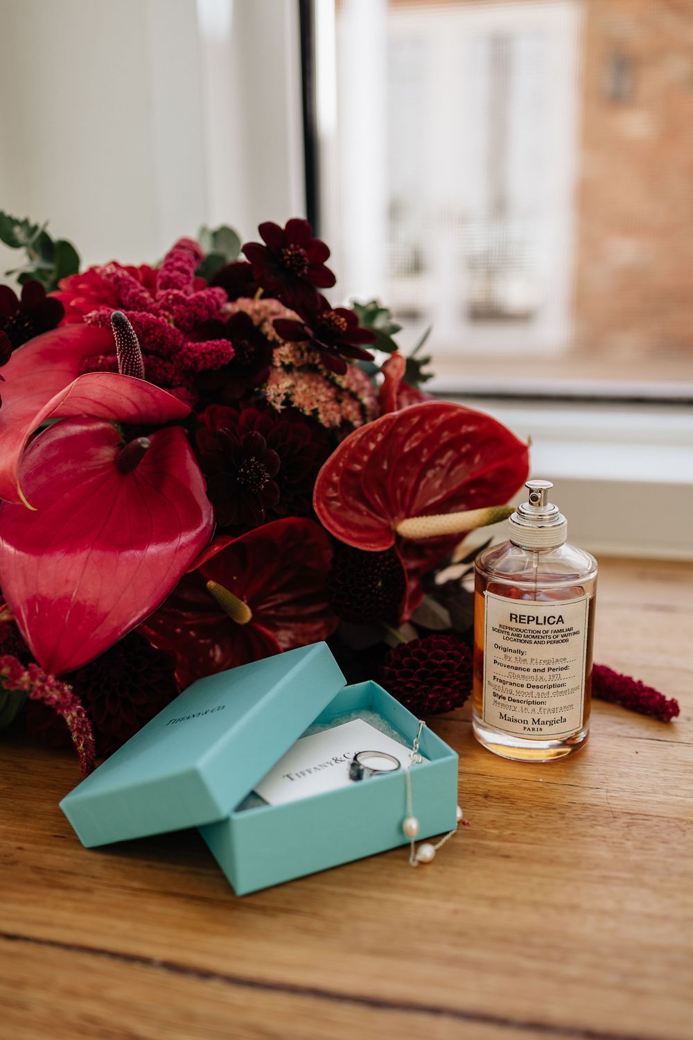 Red floral arrangement with anthurium flowers sits beside a turquoise ring box and perfume bottle on wooden surface.