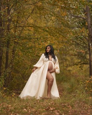 Pregnant woman in cream robe poses outdoors among autumn trees with golden foliage in a maternity photo shoot.