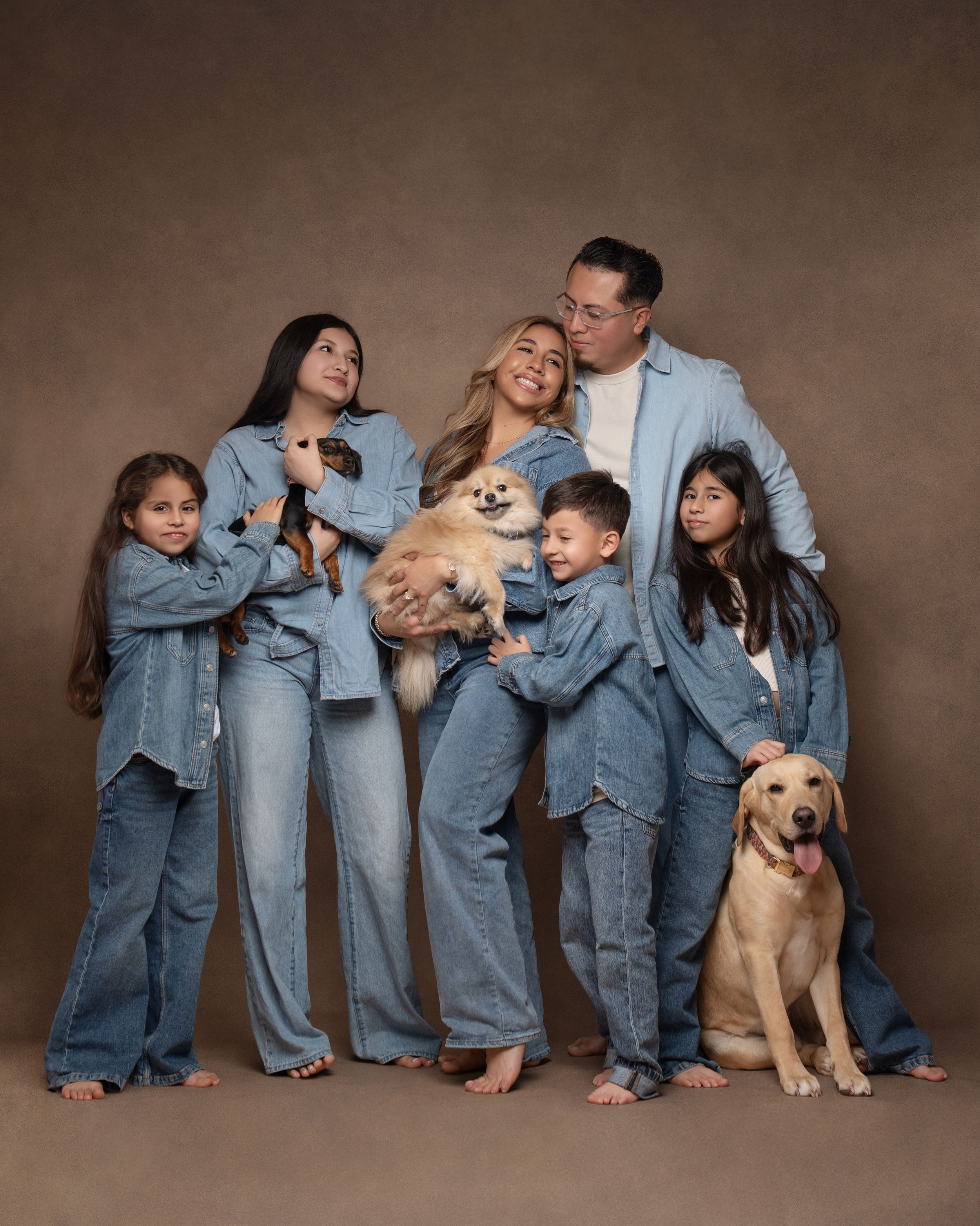 Family of five in matching denim outfits poses with two dogs including a Pomeranian and a Labrador in a studio portrait.