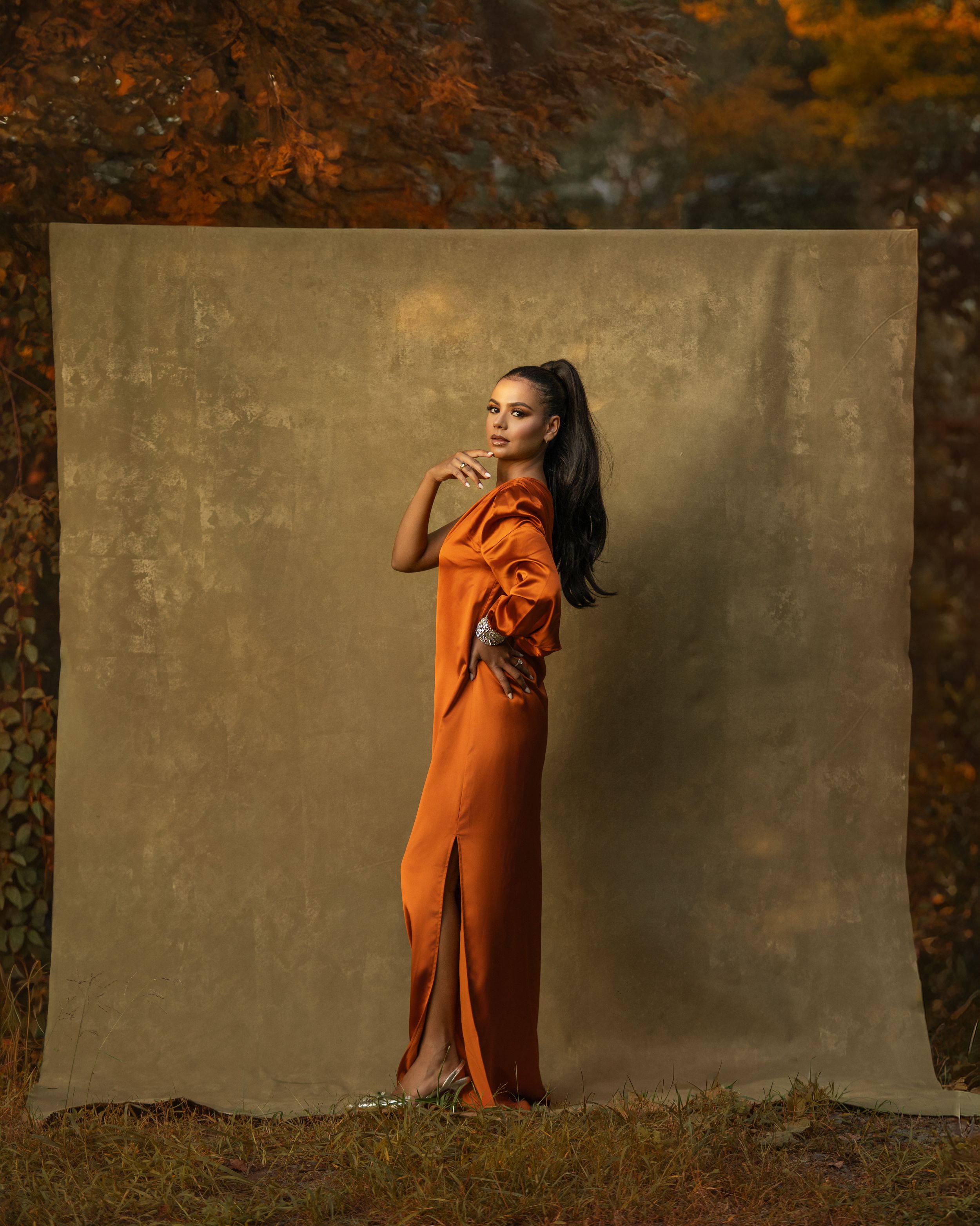 Woman with a high ponytail poses in a flowing copper satin gown against a textured olive backdrop in an autumn woodland setting.