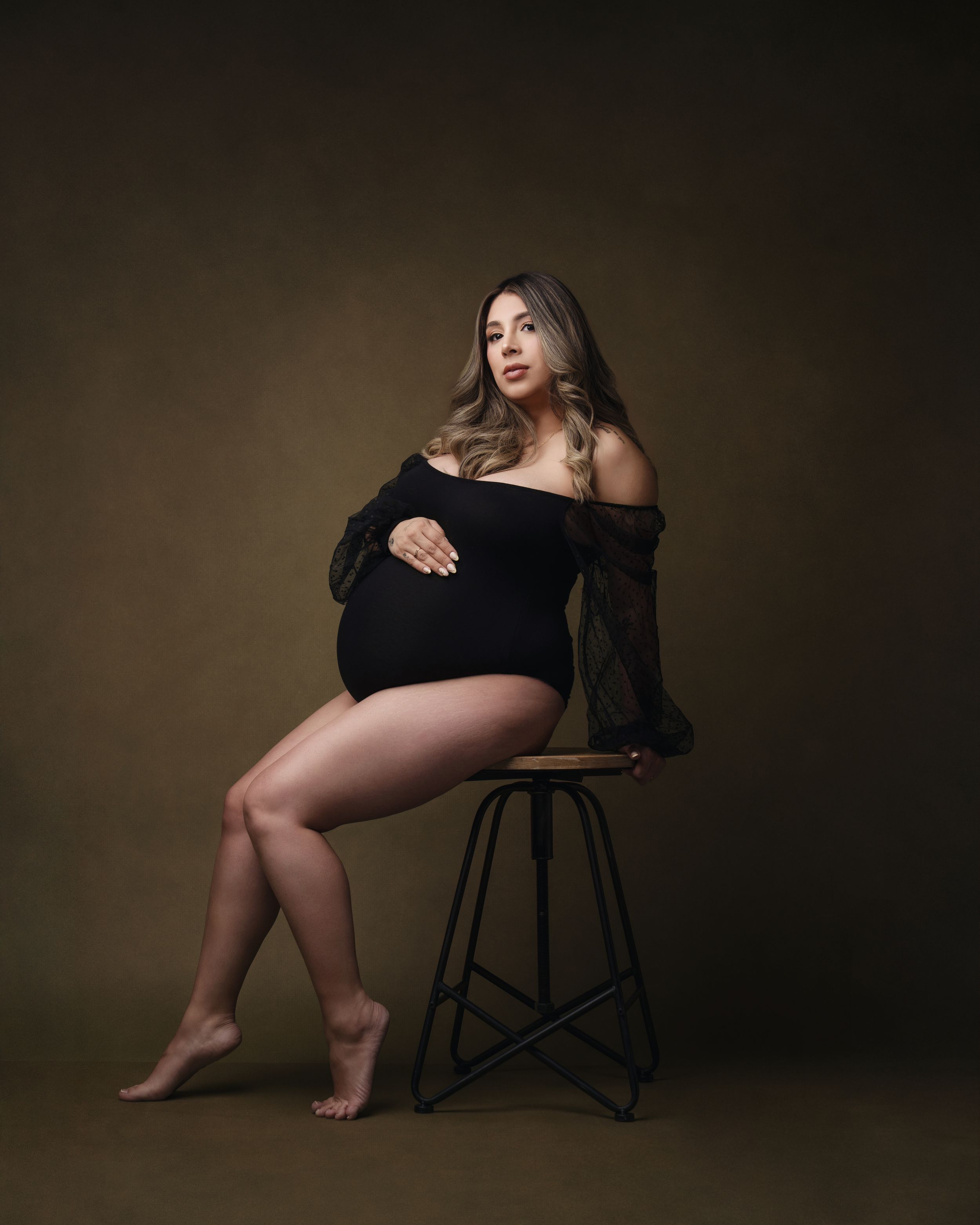 Pregnant woman in black off-shoulder bodysuit seated on stool, cradling baby bump in elegant maternity studio portrait.