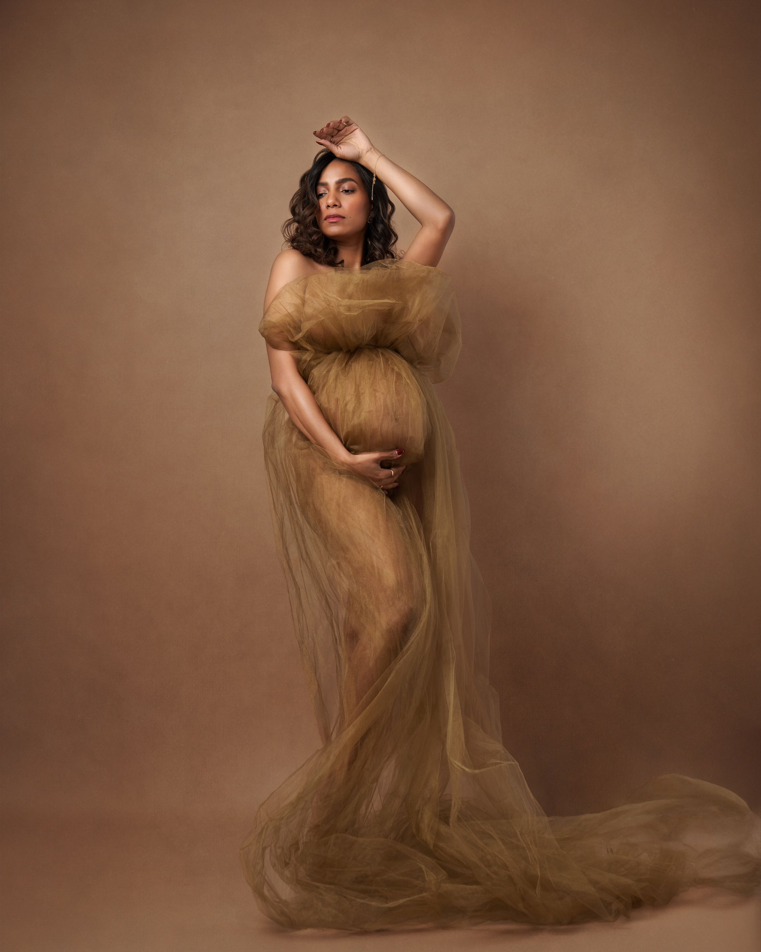Pregnant woman in flowing golden tulle gown poses gracefully against warm brown backdrop in elegant maternity portrait.