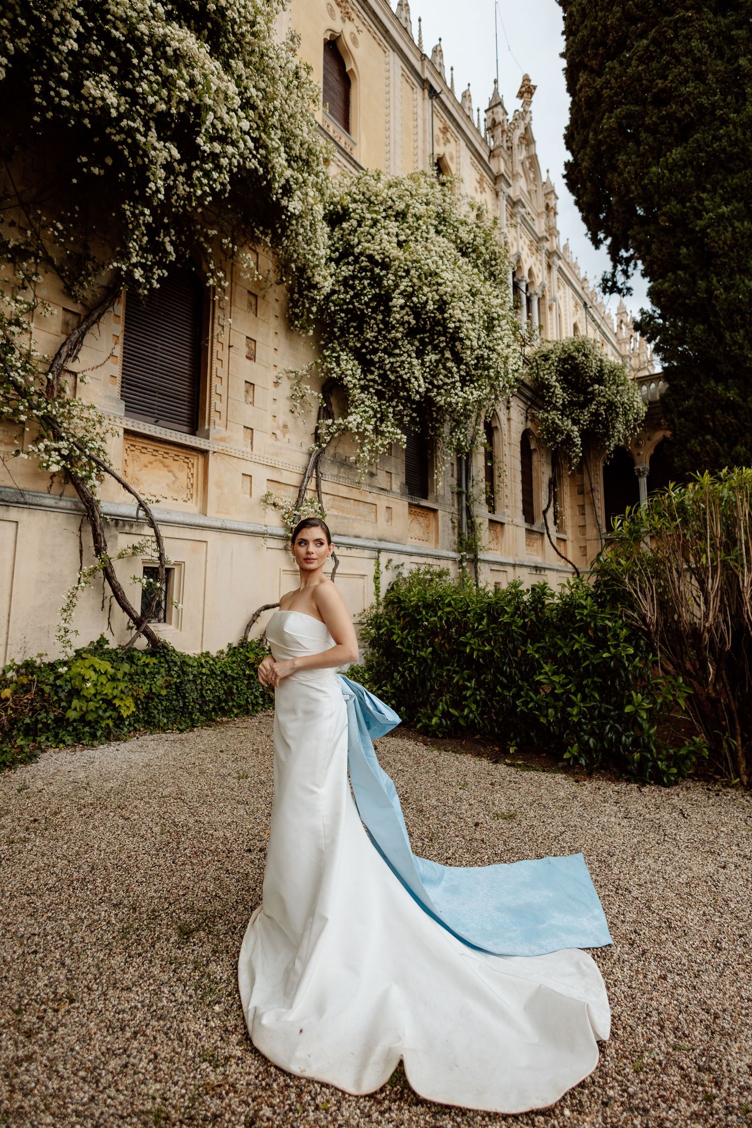 Elegant white wedding gown with light blue train detail photographed in front of historic European building with climbing vines.