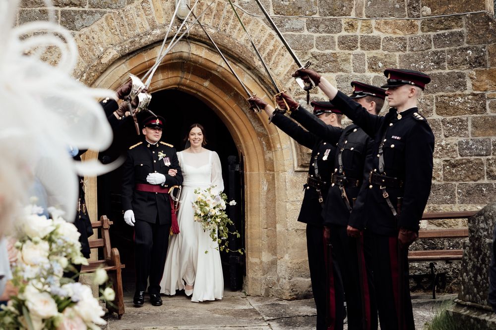 Military officers in formal dress uniform create traditional saber arch outside stone church archway during wedding ceremony.